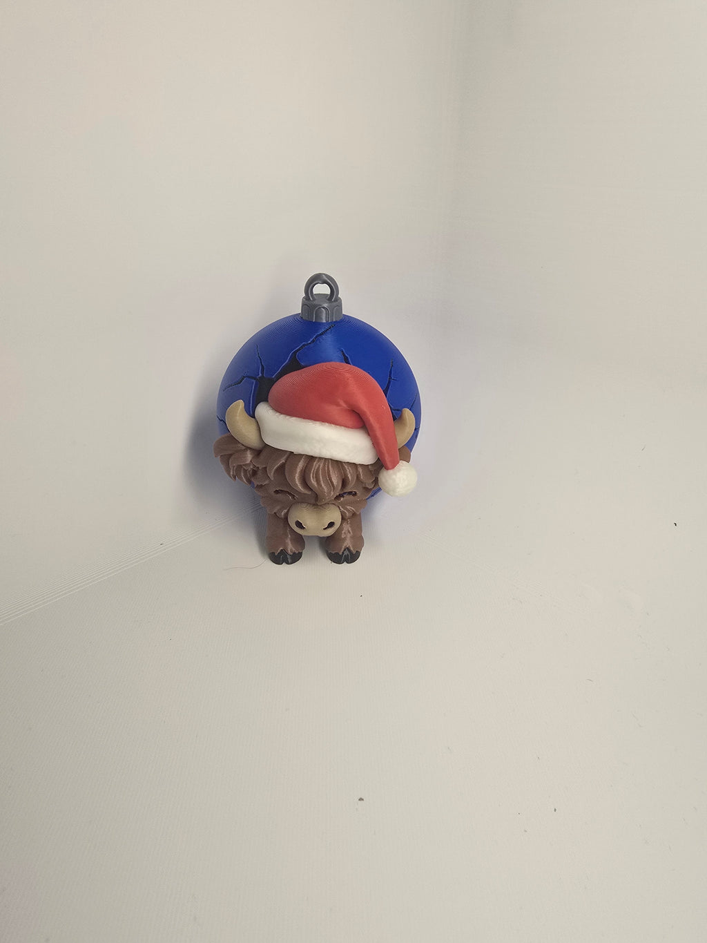 Brown Highland cow in a red Santa hat against a blue Christmas bauble-style backdrop with a hanging loop
