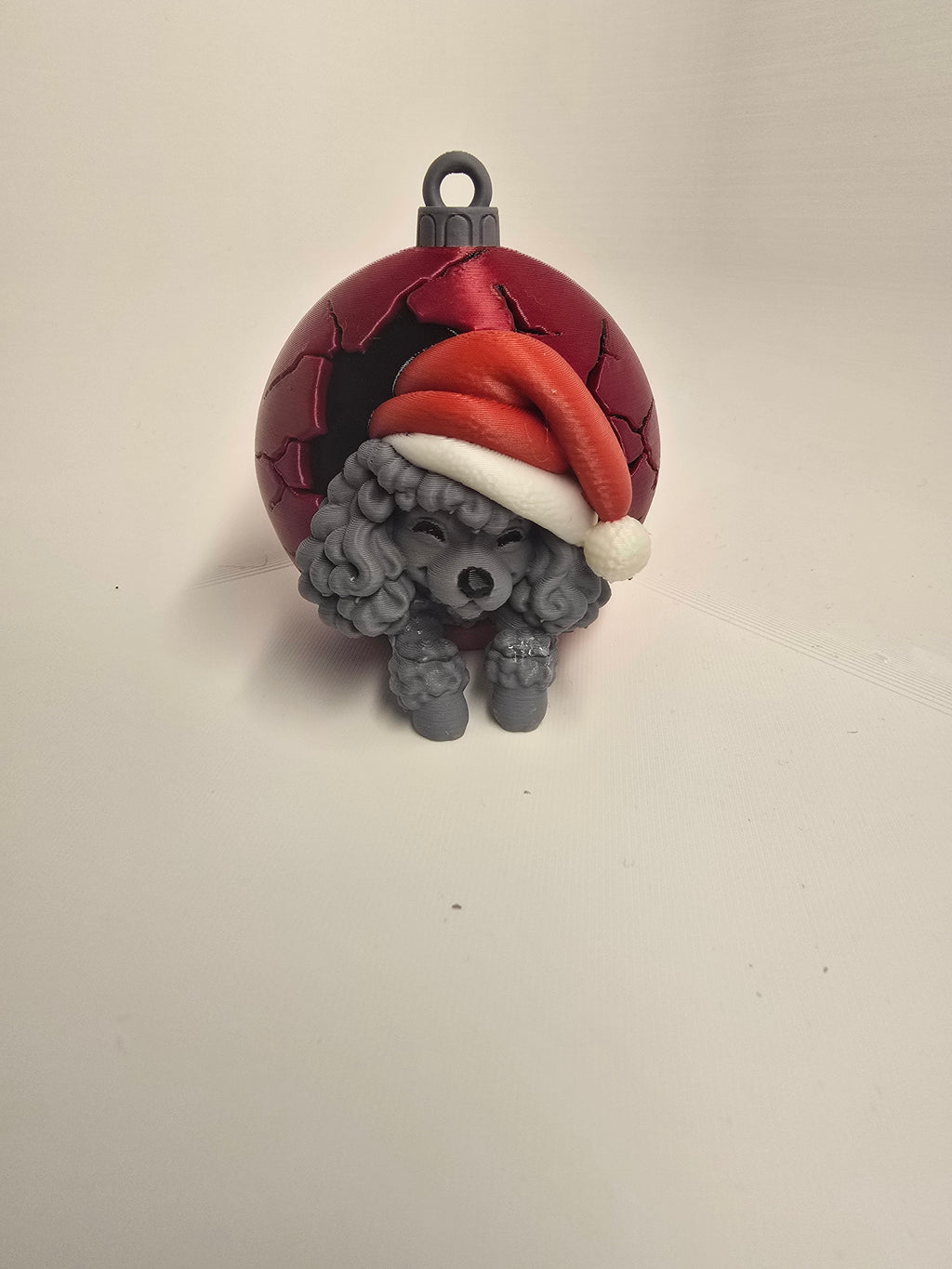 Gray curly-coated poodle wearing a red Santa hat in a red Christmas bauble-style ornament with a hanging loop