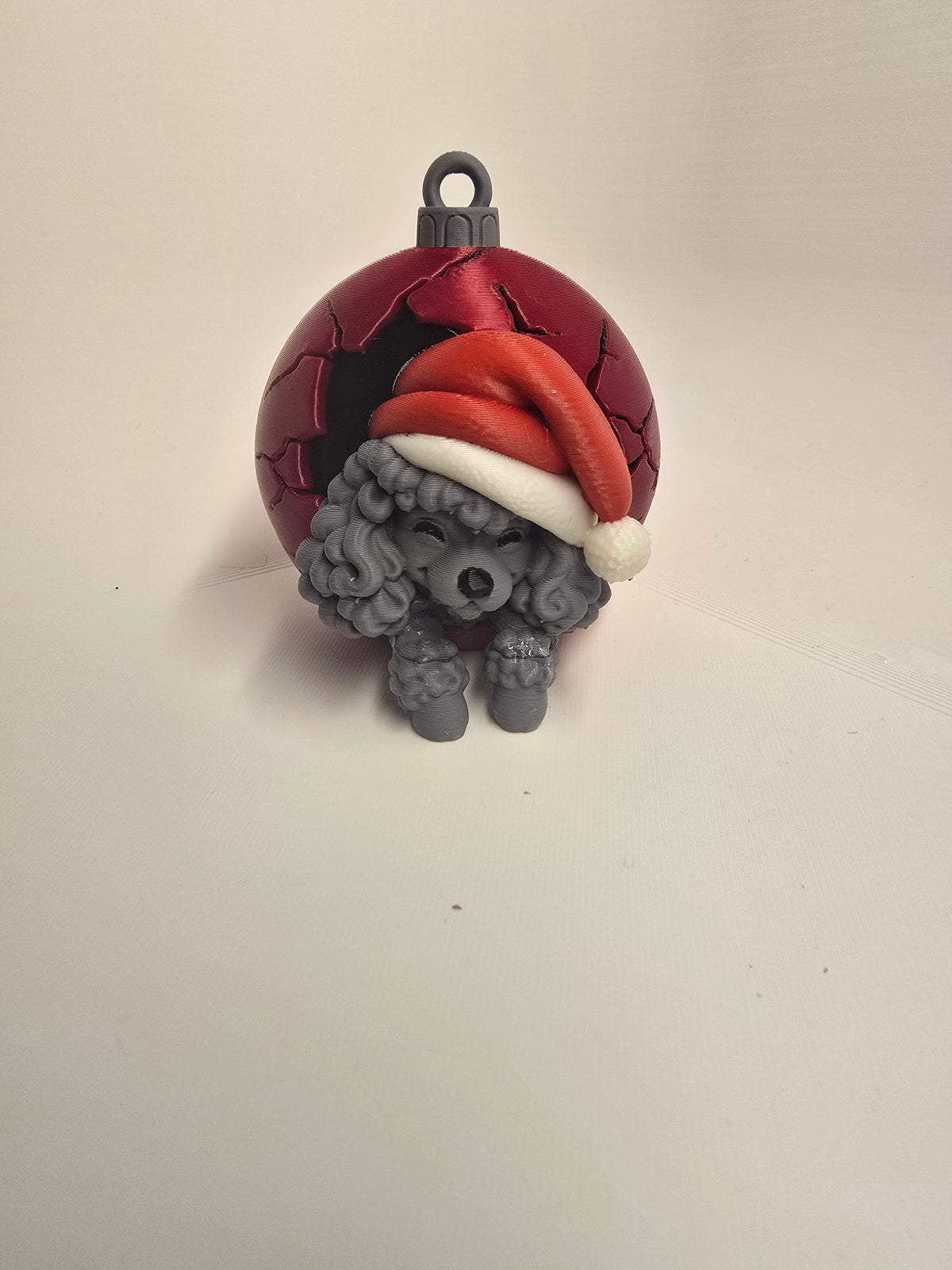 Gray curly-coated poodle wearing a red Santa hat in a red Christmas bauble-style ornament with a hanging loop