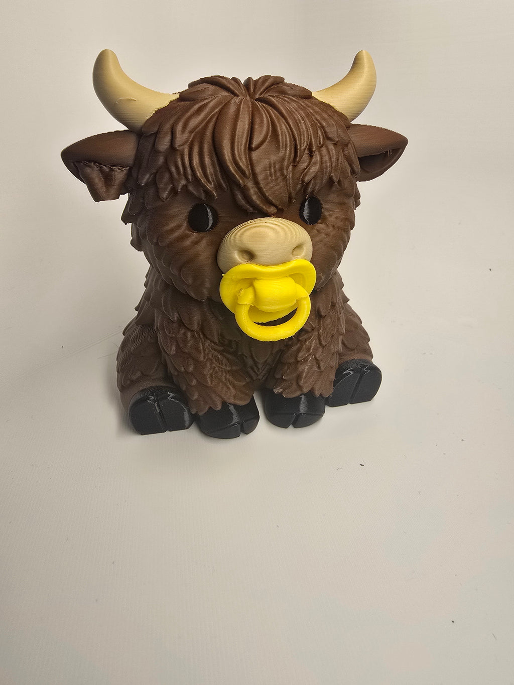 Seated baby Highland cow figurine with shaggy brown bangs, cream horns, dark hooves, and a yellow pacifier