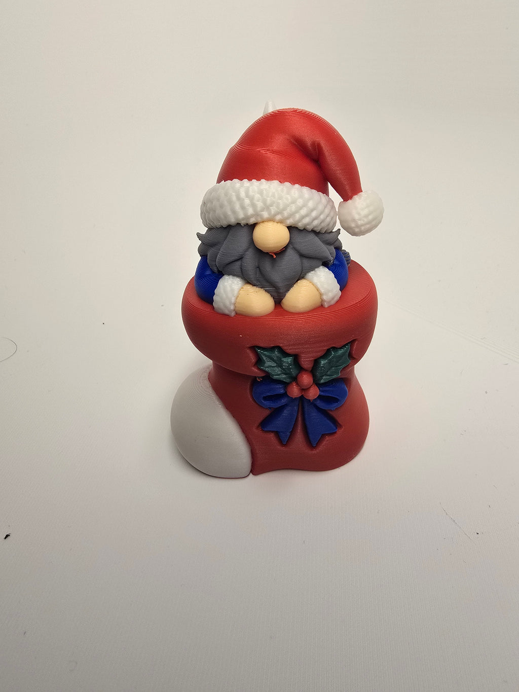 Gnome with a red Santa hat and gray beard tucked inside a red Christmas stocking decorated with holly and a blue bow